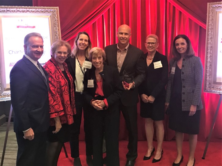 Group of seven professionals in business attire posing with an award on a red curtained backdrop.