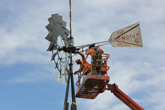 Two workers in orange safety vests repair a vintage windmill on a raised platform.