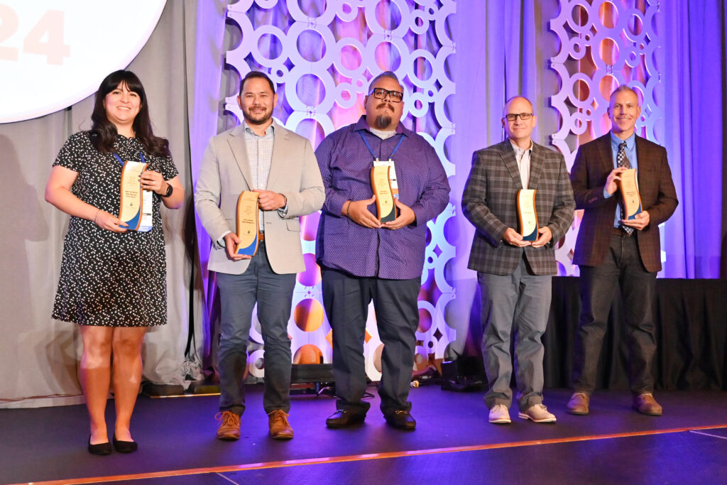 Five award recipients stand on a stage holding plaques, posing for a group photo at an awards ceremony.