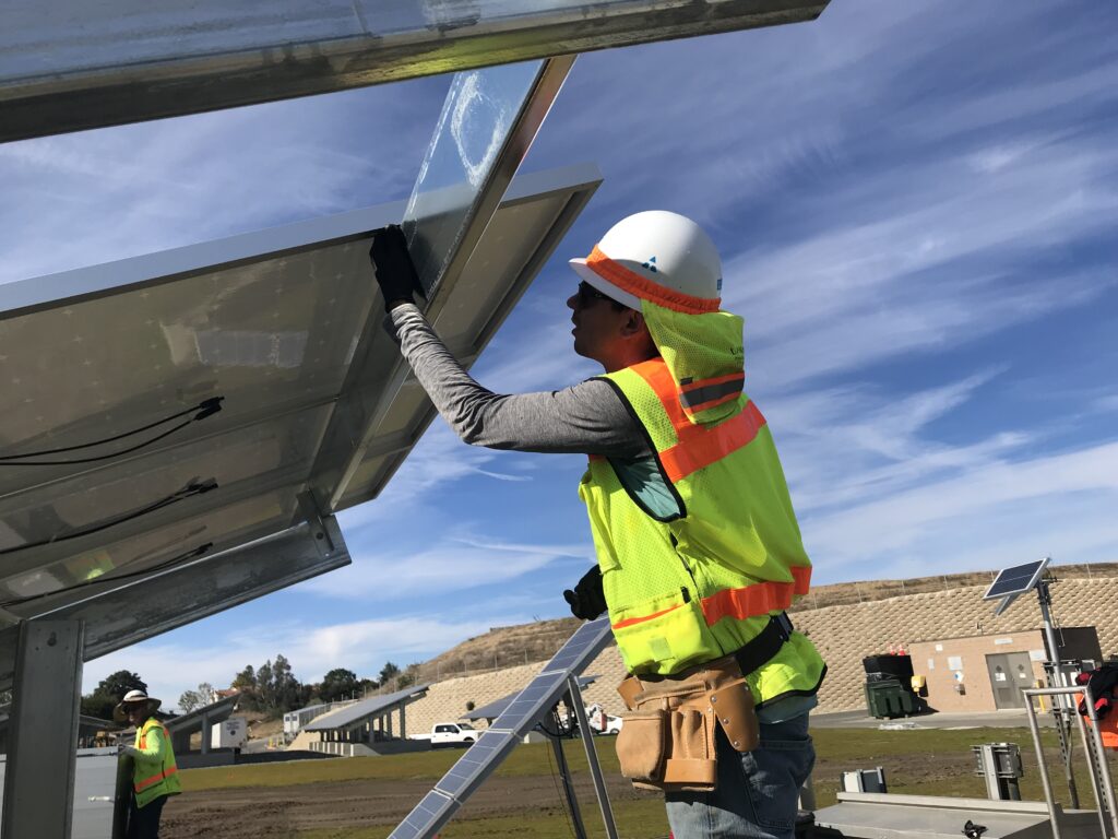 Construction worker in a hi-vis vest and hard hat installs a solar panel on a metal frame.
