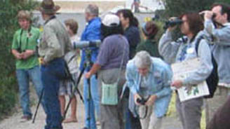 Group of people outdoors along a park path, some with cameras and binoculars, listening to a guide.