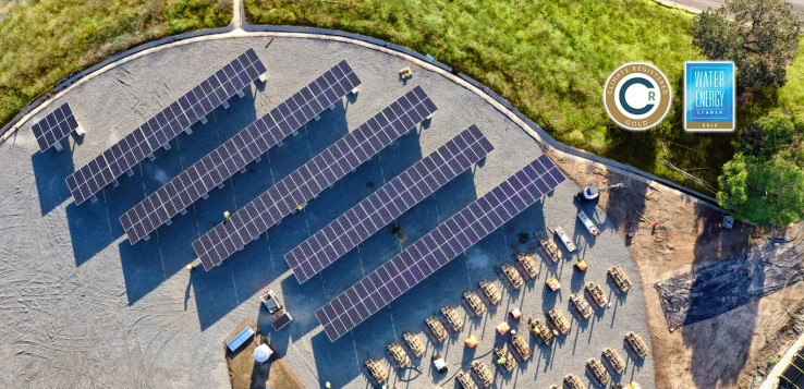 Aerial view of a solar farm with rows of solar panels on a gravel surface near trees.