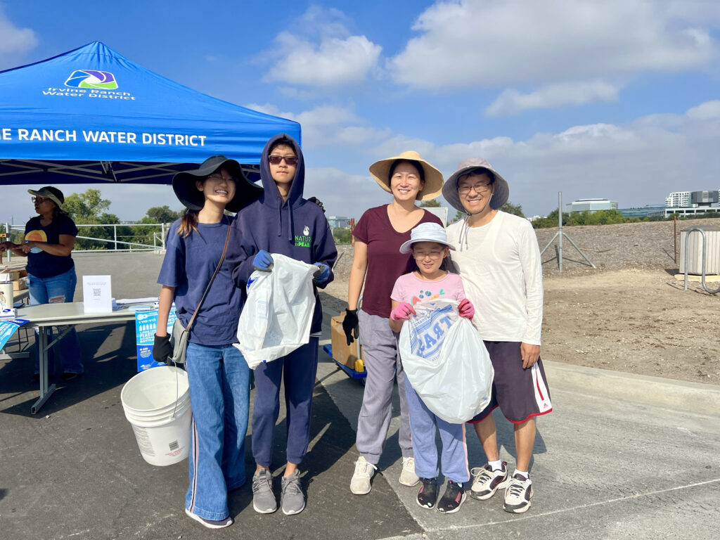 Volunteer group at outdoor cleanup under a blue Irvine Ranch Water District canopy, holding trash bags.