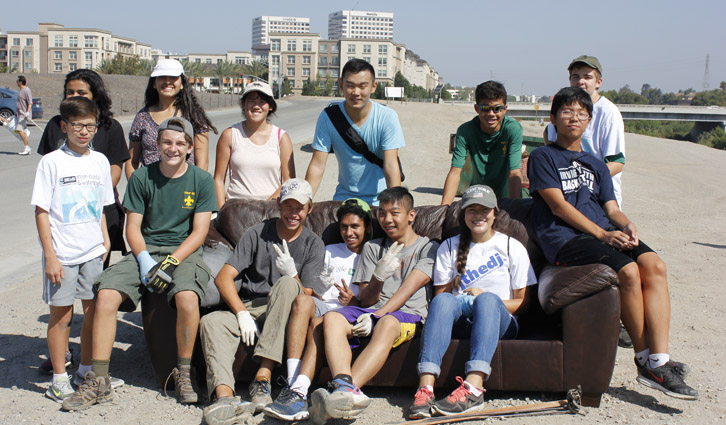 Group of diverse teens and a few adults posing on a brown couch outdoors with buildings in the background.