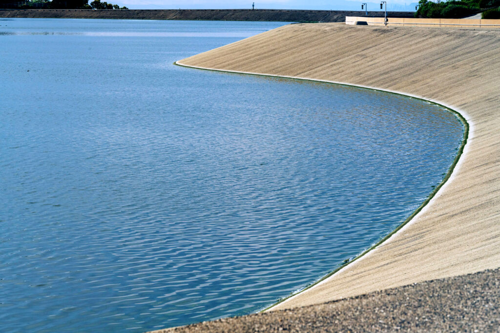 Curved concrete embankment along a blue reservoir with a distant road and railing.