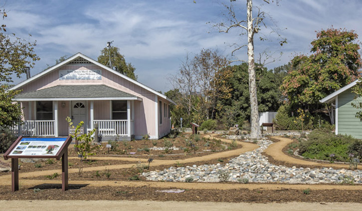 Pink wooden cottage with a small front porch, nearby signboard, and a winding pebble path through a landscaped park.