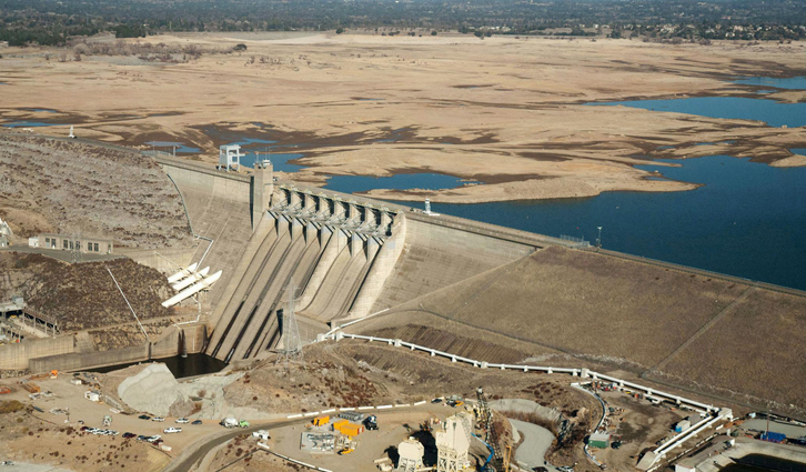 Concrete dam with cascading spillways across a river, set in a dry, hilly landscape with a blue reservoir nearby.