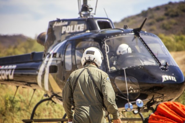 Uniformed rescuer in green suit and white helmet stands before a black police helicopter in a dry, hilly landscape.