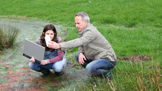 Older man and young woman crouch on a grassy field examining a laptop.