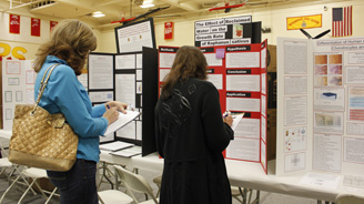 Two attendees study science fair posters displayed on red-and-white boards, taking notes.