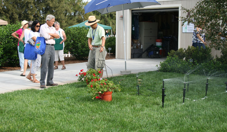 Group of adults watches a man in a straw hat watering a potted plant with a sprinkler outside a building.