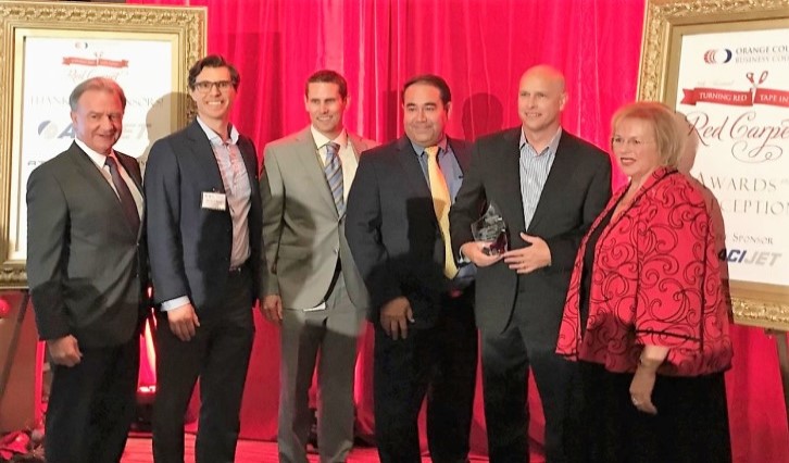 Six professionals in suits posing on a red stage at an awards ceremony; one person holds a glass trophy.