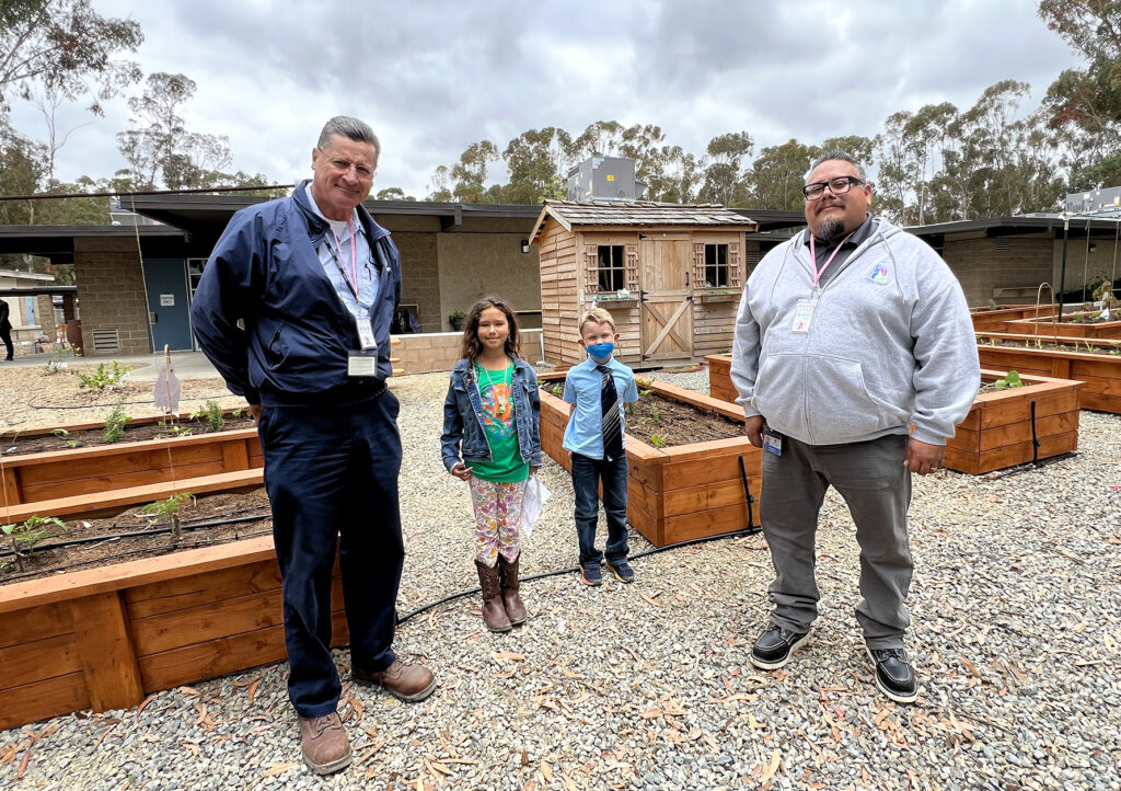 Group of four—two adults and two children—in a school garden with raised wooden beds and a shed.