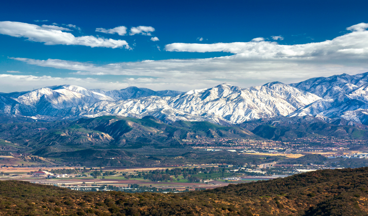 Snow-covered mountain range over a valley with fields and a distant town under a blue sky.