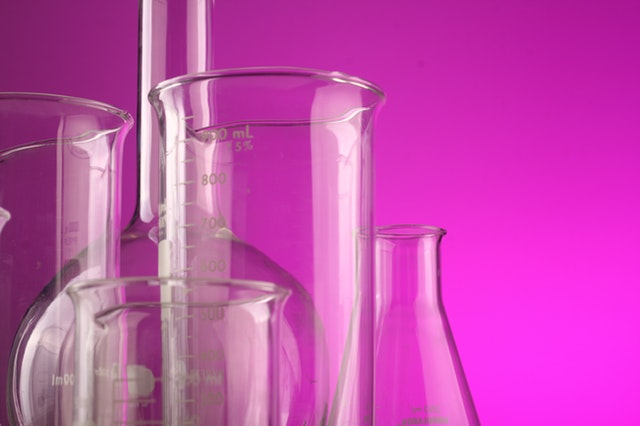 Laboratory glassware including beakers and a graduated cylinder against a vibrant pink background