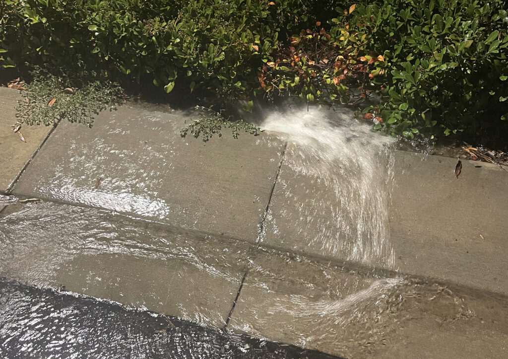 Water spills over a curb onto a sidewalk beside a green hedge, creating a small flood.