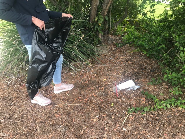 Person in a dark jacket and blue jeans collects litter with a black trash bag in a garden bed; a plastic bottle lies on mulch nearby.