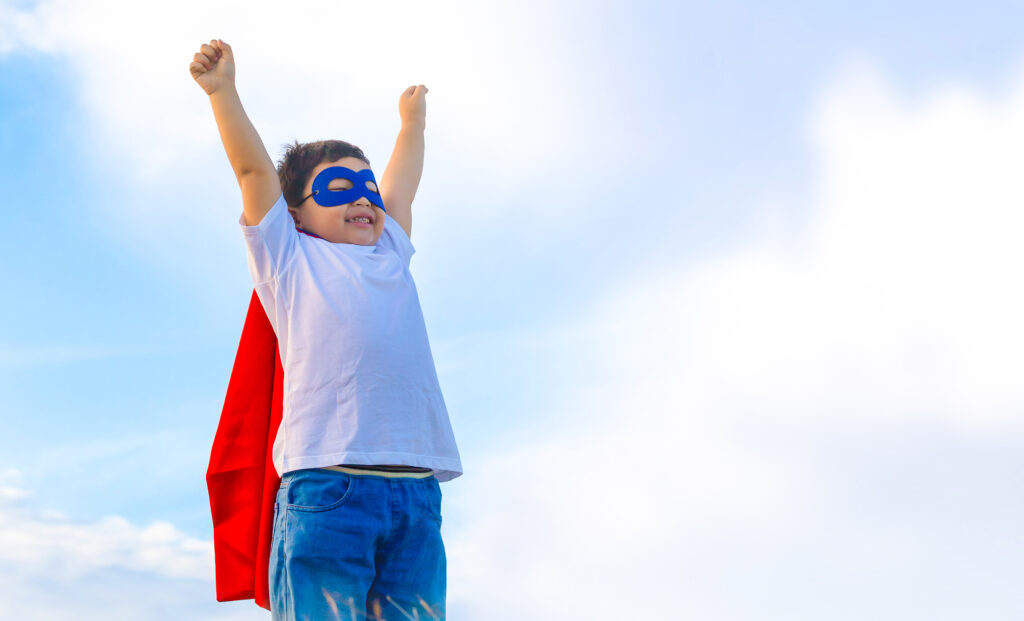 Young child in a blue cape and mask triumphantly raises arms against a bright blue sky.