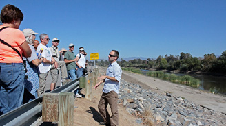 Tour guide explains riverbank project to a group of hikers beside a rocky embankment under a blue sky.