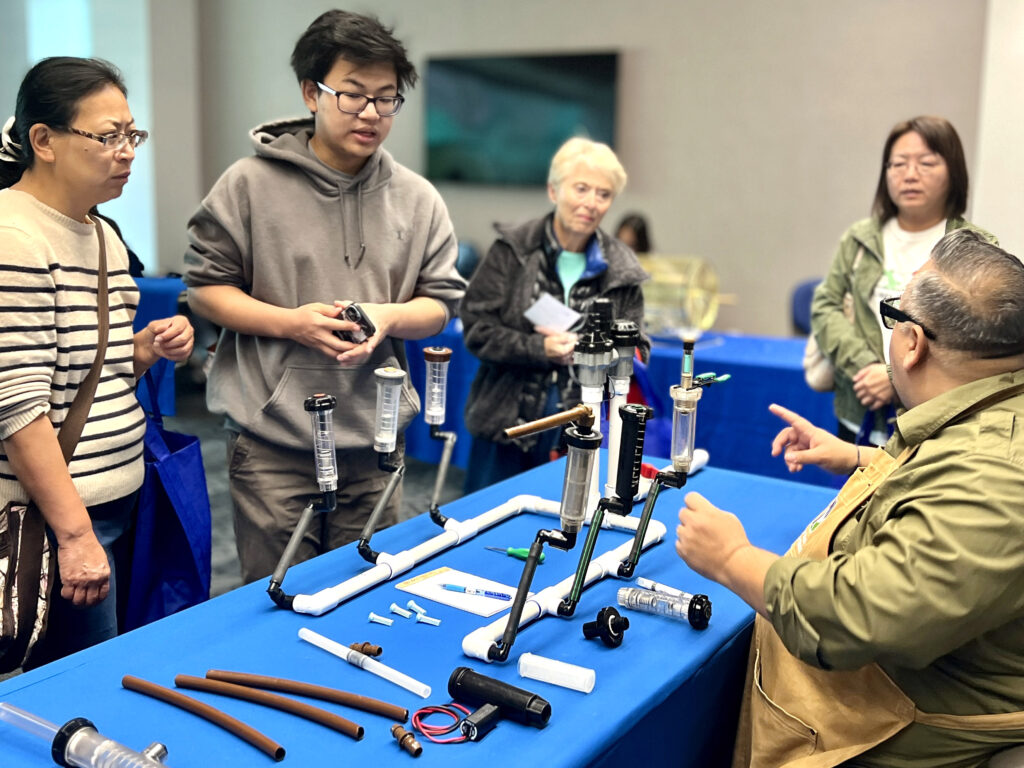 Instructor explains a pipe-based water filtration setup to a small group gathered around a blue table.