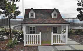 Small white porch-front garden shed with brown shingled roof and two dormer windows.